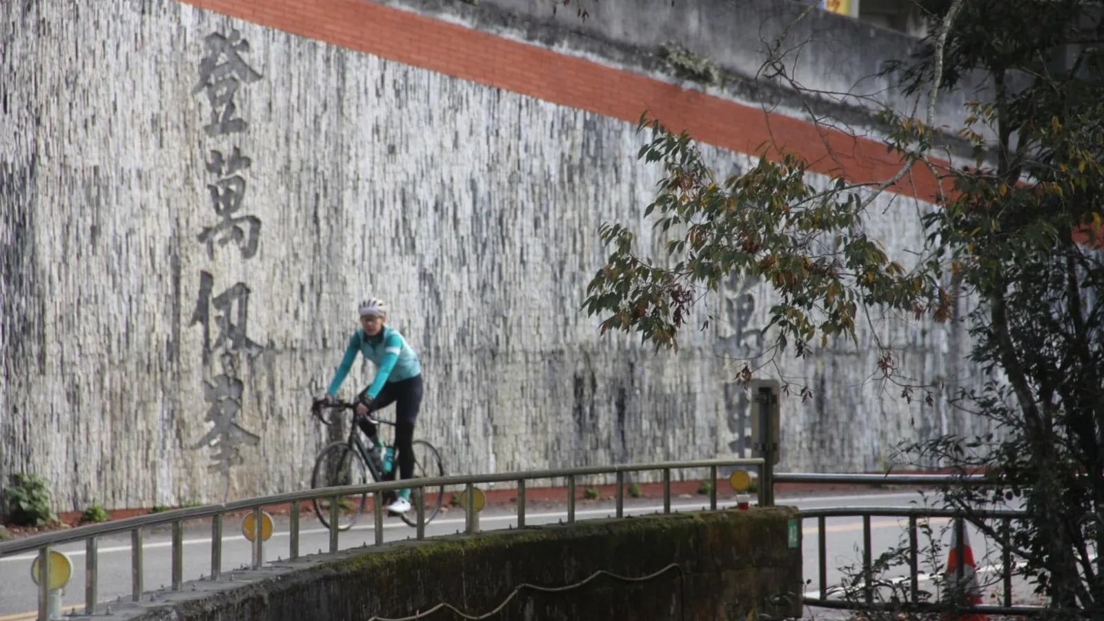 公路車騎士通過中橫公路沿線的橋段，背景是刻字的擋土牆
